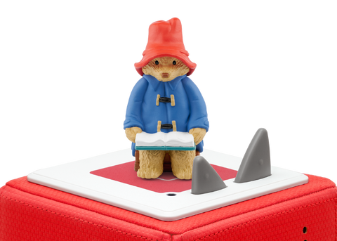 Vorderansicht der TONIES Figur Paddington auf rotem Case; Paddington in blauem Mantel und rotem Hut, geöffnetes Buch.